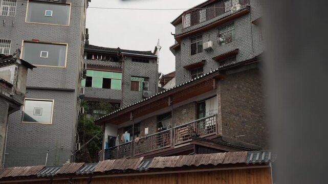 Weathered balcony apartments with hanging laundry, narrow alley and brick facades, rusted railings, satellite dishes, worn tiles and damp plaster, resident tending potted plants on balcony, overcast