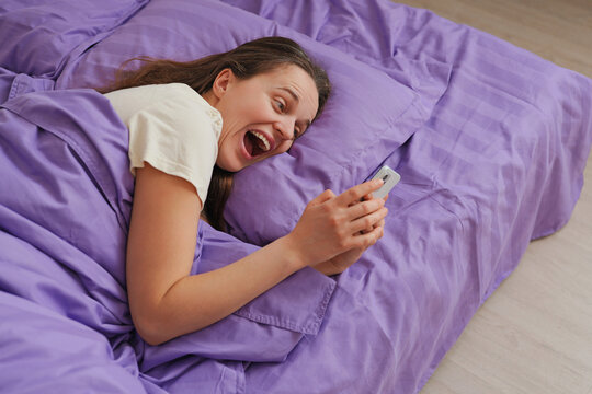 A young woman relaxes in a bed covered with purple bedding. She smiles while looking at her smartphone, enjoying a moment in the morning