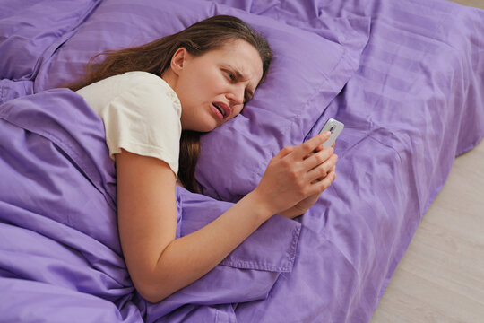 A person lies in bed wrapped in purple blankets. She looks at her phone with a concerned expression. It is evening and the room is dimly lit