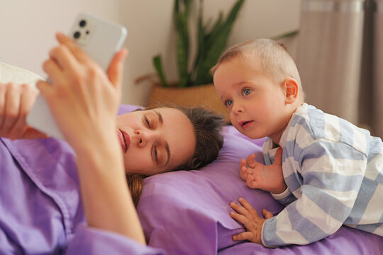 A mother lies on her side on a bed while holding her phone. Her young child leans close, looking at the phone screen. The room has simple decor and soft light