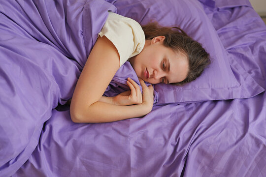 A woman lies in bed holding a pillow, looking relaxed. The bedroom features purple bedding and soft light, creating a simple and cozy scene in the daytime