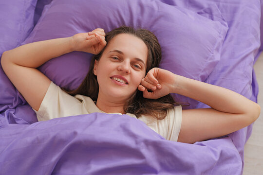 A woman lies in bed with purple sheets and pillows. She stretches her arms and smiles as sunlight filters into the cozy room in the morning