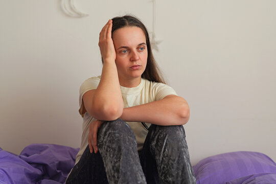 A woman sits on her bed with her hand on her head, deep in thought. The room has soft colors and the bed is made with purple bedding