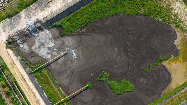 A bird's-eye view drone image of the sludge and solid waste accumulating in the wastewater treatment lagoon creates an abstract texture of pollution.