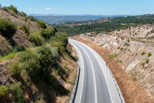 Perspetiva a&eacute;rea de uma estrada sinuosa atravessando cortes de encostas &aacute;ridas e vegeta&ccedil;&atilde;o mediterr&acirc;nea sob um c&eacute;u limpo