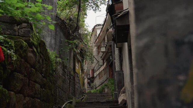Narrow mossy stone alley with steps, framed by aged stone walls and climbing stairway leading through village, hanging ivy and lantern, scattered red cloth accent, filtered daylight through leafy