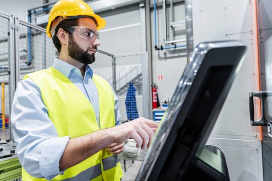 Young engineer wearing hardhat operating touch screen machine in factory