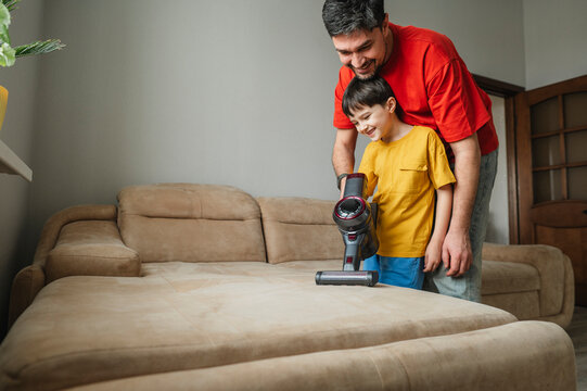 Father and son cleaning sofa together with vacuum cleaner indoors