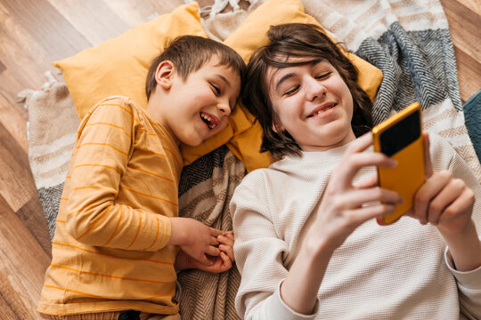 Brother and sibling lie on floor at home laughing at smartphone