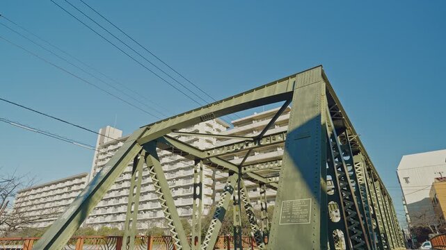 Fukuju Bridge's yellow-green steel truss fills the frame against vivid blue sky in Tokyo, with a large residential building behind, captured with a tilt-down camera movement revealing the full structu