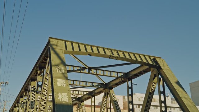 Fukuju Bridge's yellow-green steel truss fills the frame against vivid blue sky in Tokyo, with Japanese nameplate and urban buildings visible, captured as a static low-angle close-up shot.