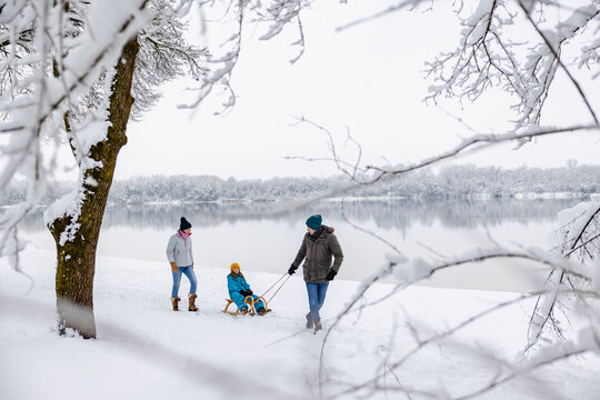 Mother and father pulling child on sled by winter lake outdoors