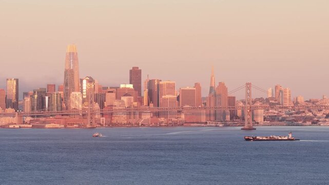 Sweeping view of the San Francisco skyline, including the Salesforce Tower and the Bay Bridge, bathed in the warm, golden light of sunrise as a cargo barge crosses the calm water