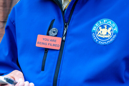 A uniformed Belfast City Council litter warden displaying a prominent warning badge stating "you are being filmed," indicating the use of body-worn surveillance cameras.