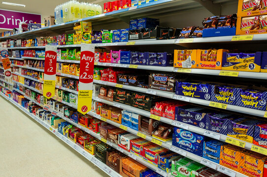 A vibrant supermarket aisle in a Tesco store featuring neatly organized shelves stocked with a wide variety of boxed chocolates, colorful candy bars, and festive sweet treats for shoppers.