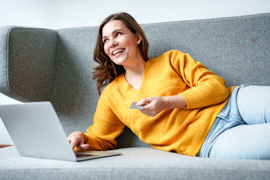 Happy woman reclining with laptop on sofa doing online shopping through credit card at home