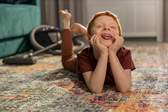 Cheerful redhead lboy ying on the carpet at home next to a vacuum