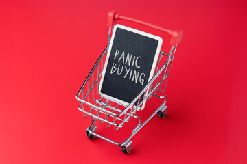 A shopping cart and a chalkboard with text PANIC BUYING against red background. Consumer hoarding, retail crises, and economic anxiety concept. © izzuan