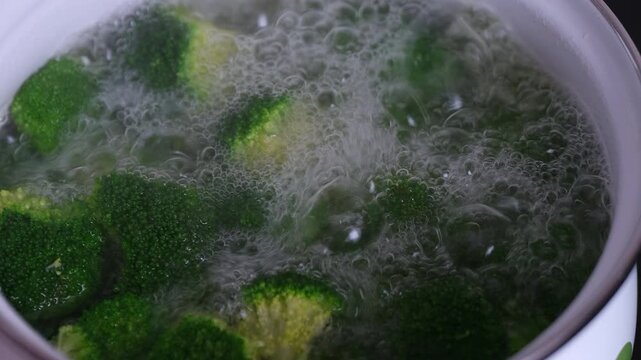 A hand lifts a glass lid from a pot of fresh broccoli cooking in boiling water. Close-up of fresh green broccoli florets boiling in a pot of hot water with rising steam.