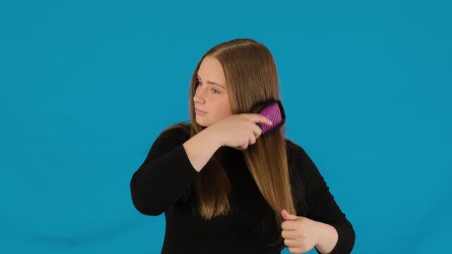 Attractive woman uses comb to brush tangled strands. Female combs hair starting from tips to minimize breakage on blue background