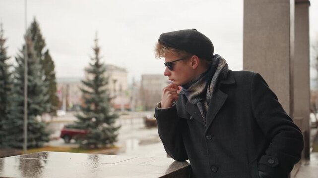 White creator leaning on wet railing, contemplative urban muse of white male creator in beret and scarf, scanning rainy plaza and architecture, cinematic editorial tone with muted palette