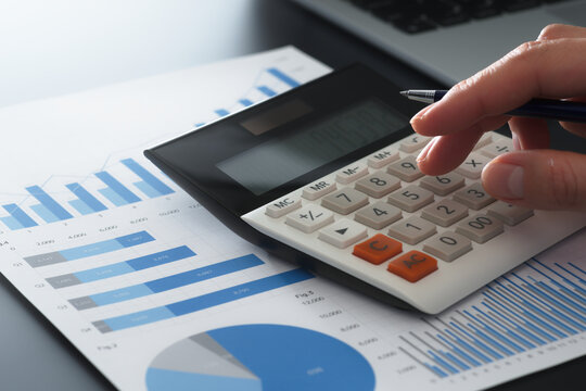 Business financial analysis and calculations. Accounting management and budget planning. Close-up of businessman's hand using calculator on report with graphs.