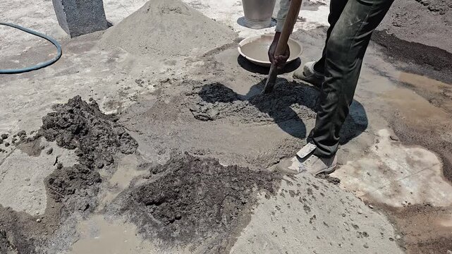 A worker manually mixes cement, concrete, & aggregate on a slab floor. Using a shovel, they pour the mixture into a bowl to reach right consistency for waterproofing or plastering a wall effectively.