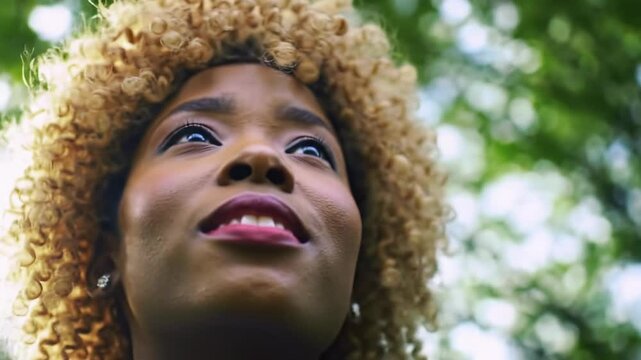 Extreme close-up video of a young Black woman with blonde-highlighted curly hair, radiant skin, serene upward gaze, golden hour glow on lush green. Tranquil optimism concept