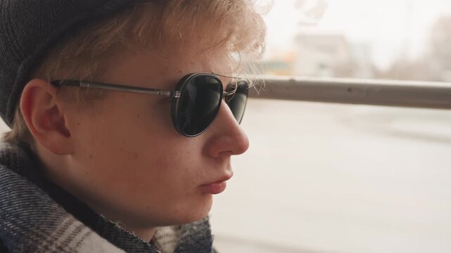 Young man staring out window, contemplative creator on urban commute, profile portrait with sunglasses and cap, soft motion blur outside, introspective cinematic mood and quiet storytelling