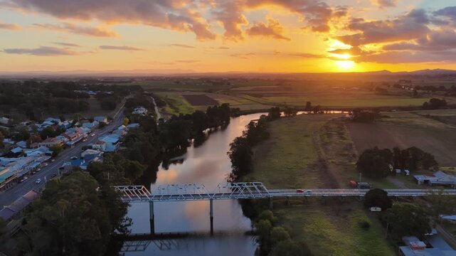 Aerial video flying over historic Morpeth bridge and along Hunter River toward colourful sunset in NSW, Australia. Scenic landscape showing river, bridge structure and warm evening light 