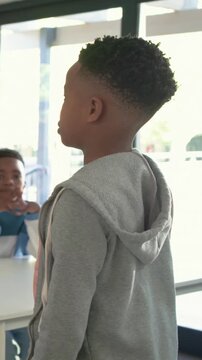 Vertical video: Seeing peer, teen in gray hoodie turning talking at class table by glass door