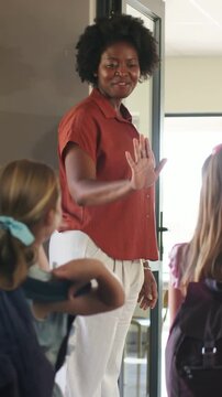 Teacher greeting arriving children at doorway, offering high-fives and guiding backpacks inside