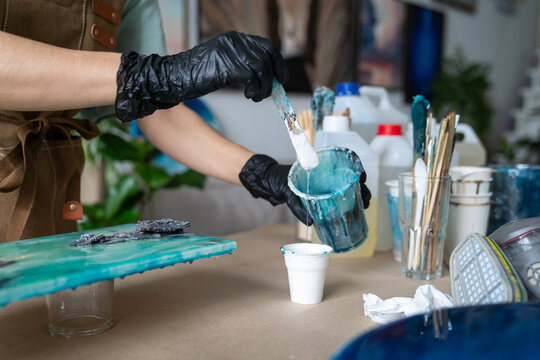 Attentive woman working with epoxy resin at worktable in art studio, carefully pouring epoxy resin in paper cup, female crafter in gloves adding varnish in cup, process of creating epoxy resin art.