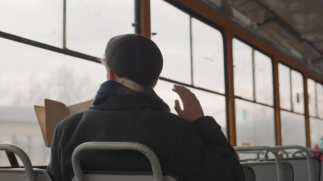 White creator on tram studying book and adjusting cap, pages open against fogged window young white researcher scratches head, flips pages and gestures while reading, capturing quiet moment of problem