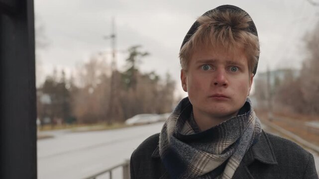 Young white creator at bus stop, overcast morning with wool cap and patterned scarf, sideways glances toward passing traffic, cold commuter atmosphere, cinematic portrait emphasizing solitude