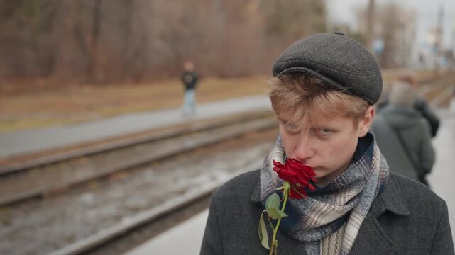 Young white man with rose on platform, defiant creator glare, wet pavement and passing commuters, wool scarf and beanie, urban tension, cinematic close portrait, subtle rain, raw expression