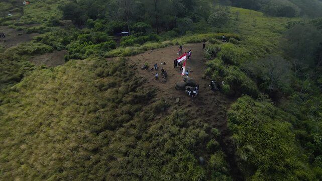 Aerial view of hikers at a grassy mountain peak with the Indonesian flag under a clear sky.