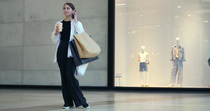 Woman talking on phone while holding coffee, standing outside boutique display, large shopping bags, professional attire suggesting busy schedule, mannequins and glossy glass backdrop