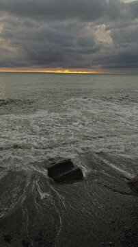 Stormy sunset over rocky shore, dark clouds with narrow golden horizon, storm chaser observation of crashing waves and foam