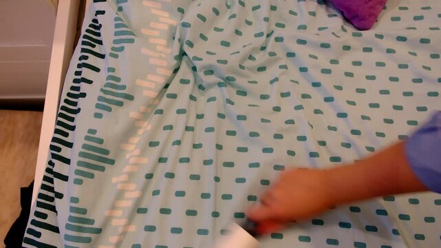 Top view of a hand rolling a lint roller across bed sheets, motion blur over a patterned fabric for a clean home routine. Removing dust and hair supports allergies control, hygiene, and comfort.
