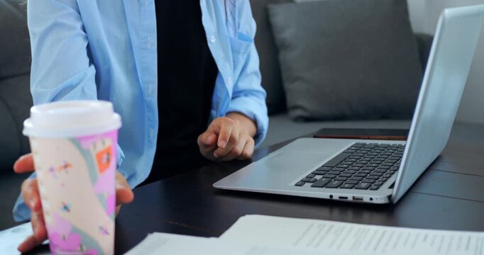 Individual reaches for takeout coffee during work. Woman balances caffeine with focus at her computer desk. Woman simultaneously types and takes coffee break at her workspace in closeup