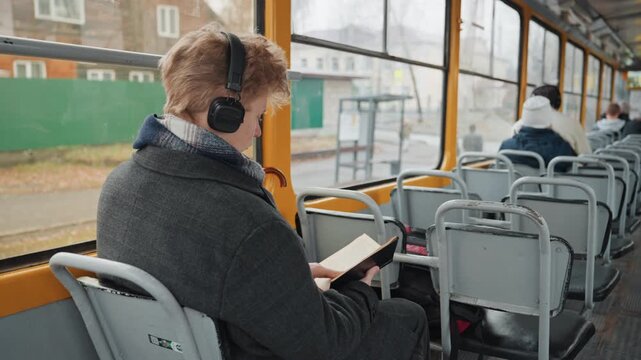 White man student creator studying on tram headphones, taking notes from book, backpack at feet, city blocks sliding by outside window, diligent focused posture, commuter research routine