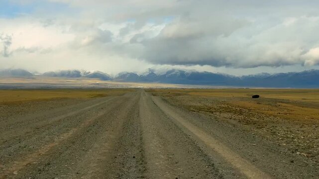Gravel road stretching from Altai towards the Mongolian border with snowy mountain ridge. Desert steppe landscape under stormy cloudy sky showcasing wild remote terrain