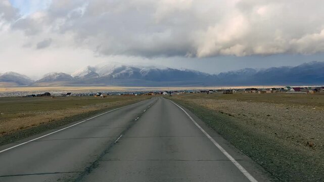 Gravel road stretching from Altai towards the Mongolian border with snowy mountain ridge. Desert steppe landscape under stormy cloudy sky showcasing wild remote terrain