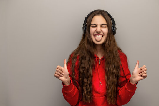 Playful Teenage Girl Wearing Headphones. Teenage girl in a red hoodie wears headphones, sticks out her tongue, and gives two thumbs up playfully.
