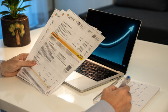 Milan, Italy 3/22/2026 Man holding Italian Iren energy utility bill over office desk with laptop screen showing upward glowing arrow chart representing rising electricity and gas market prices