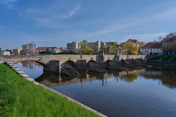 Fototapeta premium Pisek, Czech Republic - April 18, 2026 - The Stone Bridge. Historic stone bridge arches over a calm river with city buildings in the background