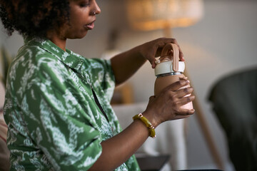 Naklejka premium Black young adult woman opening reusable water bottle at home, supporting daily hydration and sustainable routine