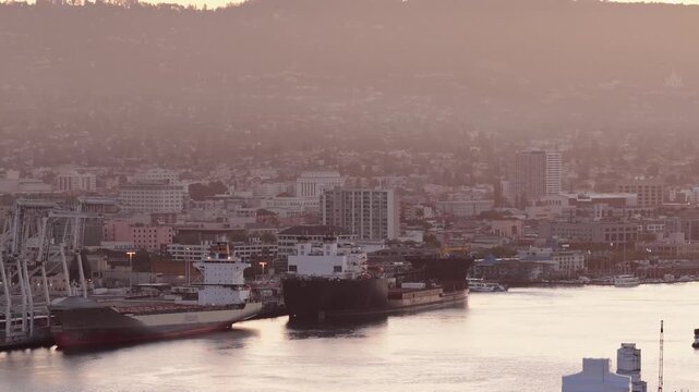 Aerial view of the port of Oakland, California, with cargo ships docked at the terminal and the city skyline and hills in the background during a beautiful, hazy sunrise over the San Francisco Bay