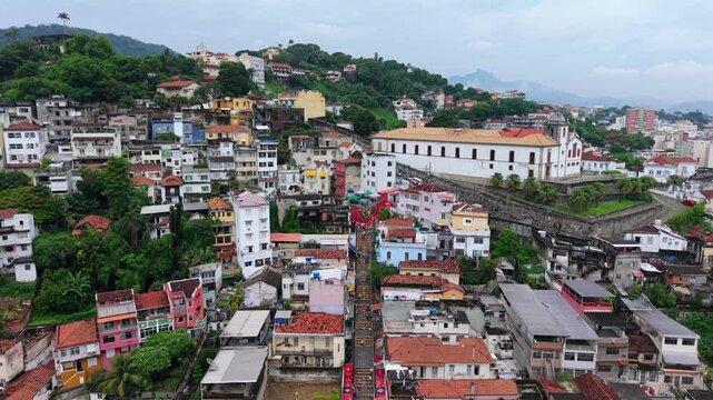 Colorful neighborhood houses at Escadaria Selar&oacute;n, Rio de Janeiro, showing vibrant mosaic steps between tropical hills and urban architecture. Pullback aerial ascent under cloudy skies.